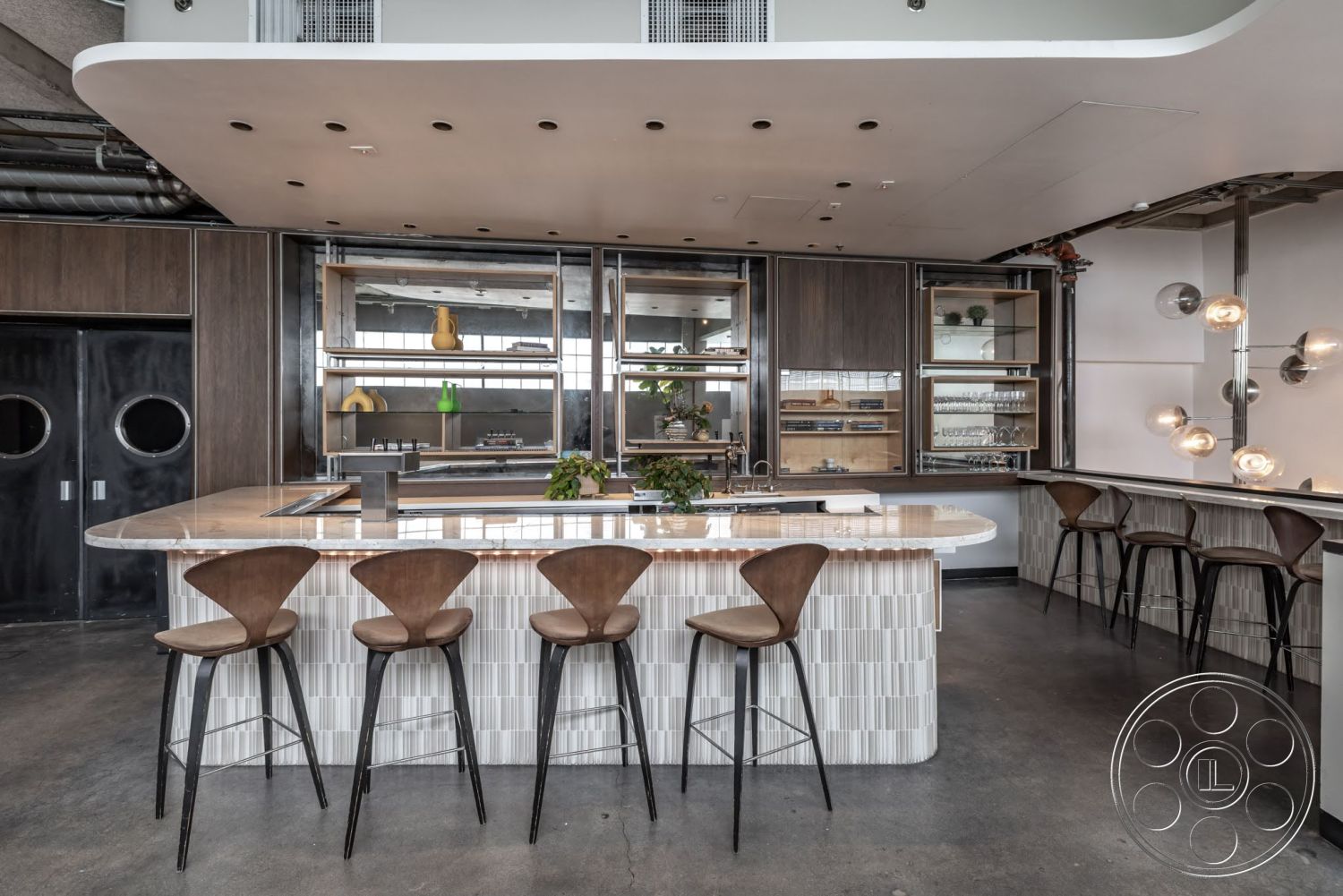 Sunset Campus - neutral color palette, wood cabinetry, concrete flooring, pendant lighting fixtures, open kitchen layout, marble countertop island, glass display shelves, bar seating with wooden stools, exposed ceiling pipes, black steel door, rounded bar counter edge