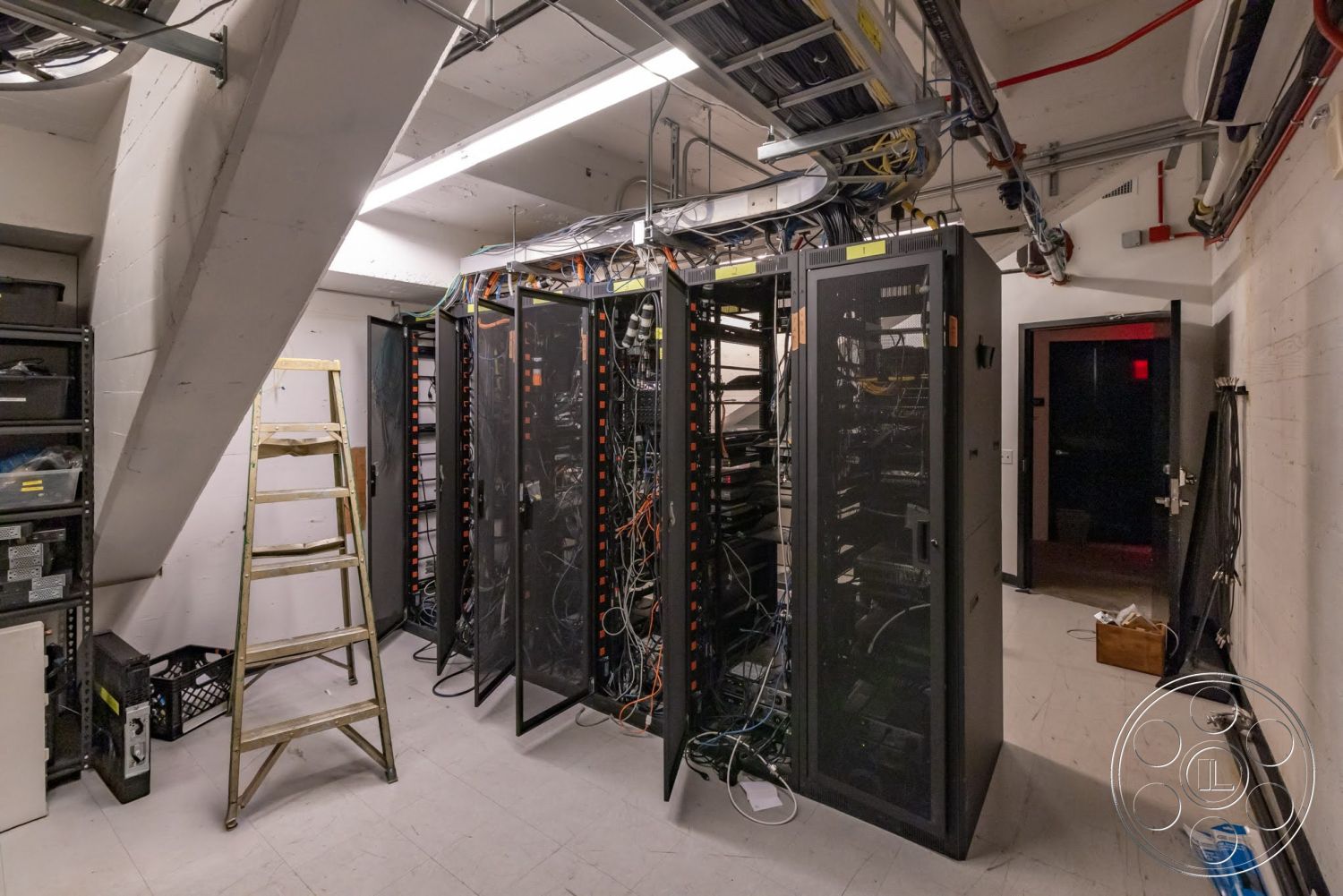Sunset Campus - interior space, exposed concrete walls, LED ceiling lights, metal server racks, fiber optic cables, stainless steel ladder, industrial ceiling infrastructure, elevated flooring, black cabinet storage, dark doorway