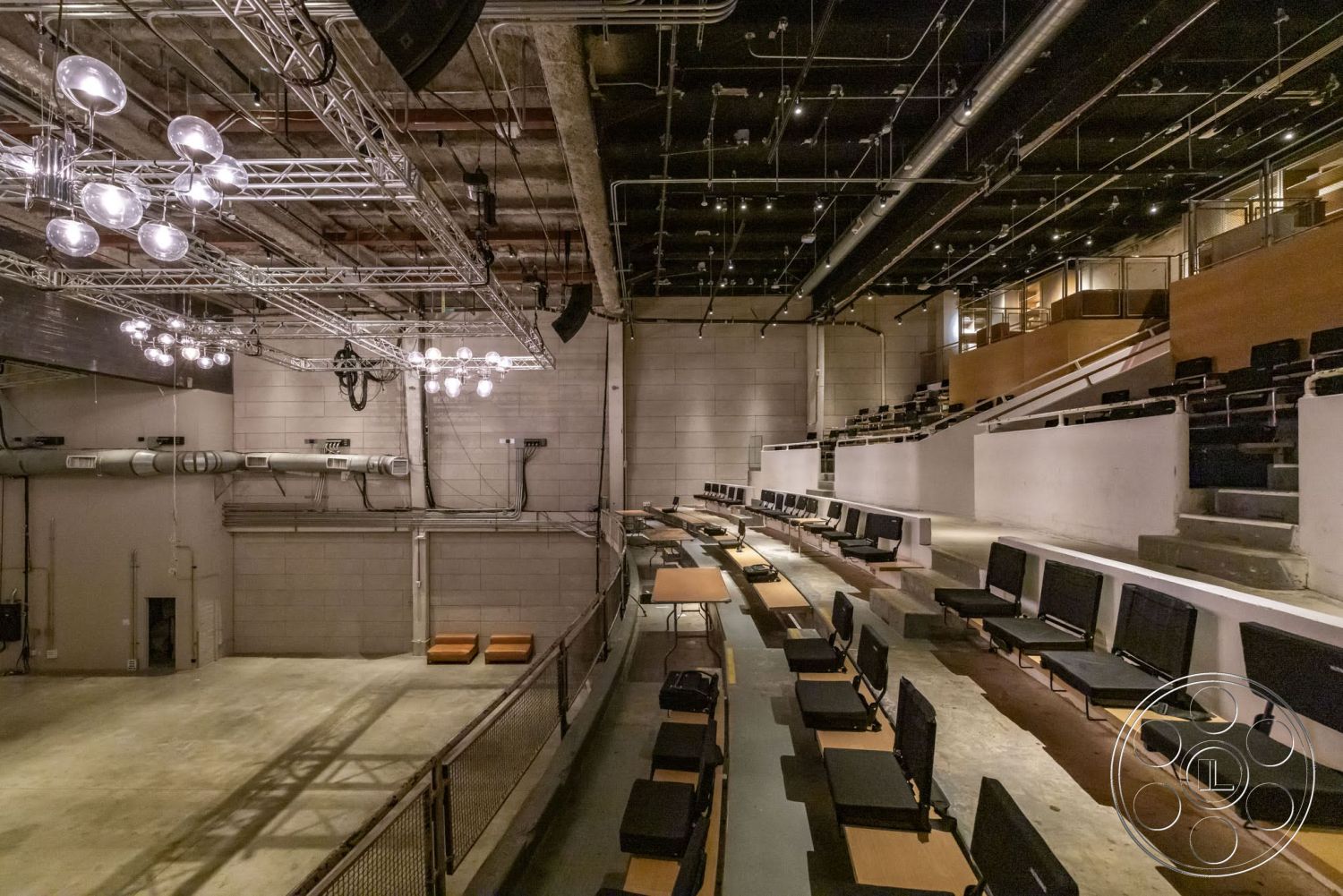 Sunset Campus - concrete floor, exposed concrete walls, curved seating arrangement, track lighting fixtures, wood paneling accents, open performance space, industrial-style seating, metal ceiling structure, multi-level audience area, industrial theater interior