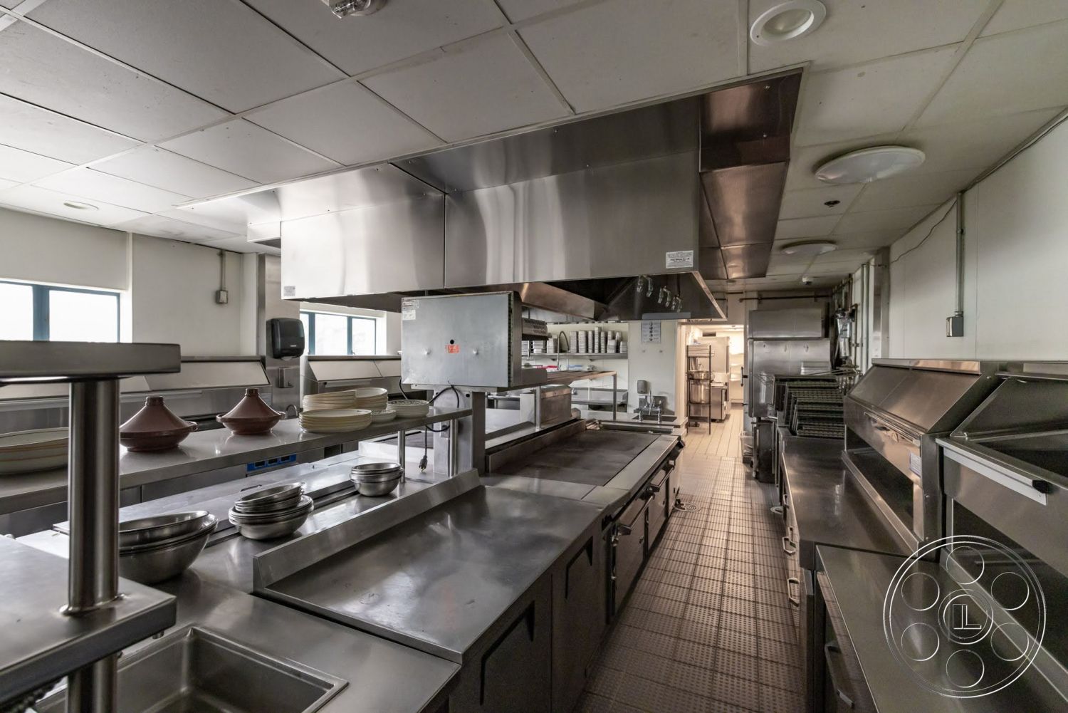 Sunset Campus - floor-to-ceiling windows, chef's kitchen, overhead lighting fixtures, modern kitchen design, stainless steel range hood, white painted walls, vinyl tile flooring, stainless steel countertops, industrial kitchen layout, commercial kitchen space, stainless steel kitchen equipment, open kitchen workspace