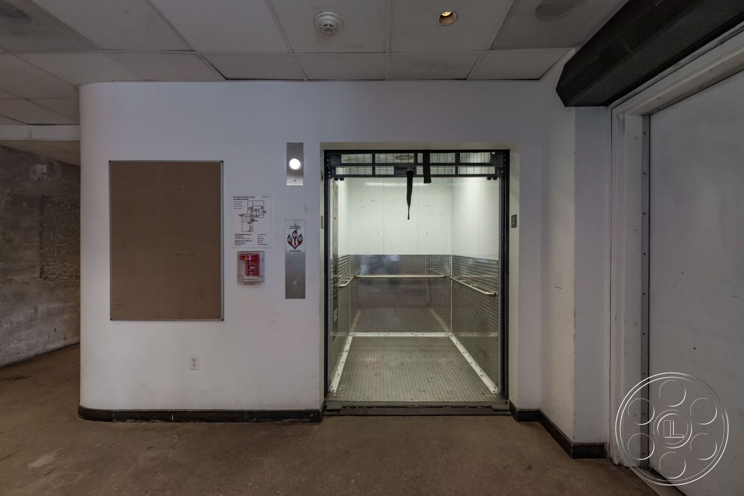 Sunset Campus - exposed concrete walls, metal elevator doors, textured concrete floor, interior elevator entrance, bulletin board wall, fire alarm panel, stainless steel elevator interior, ceiling mounted lights