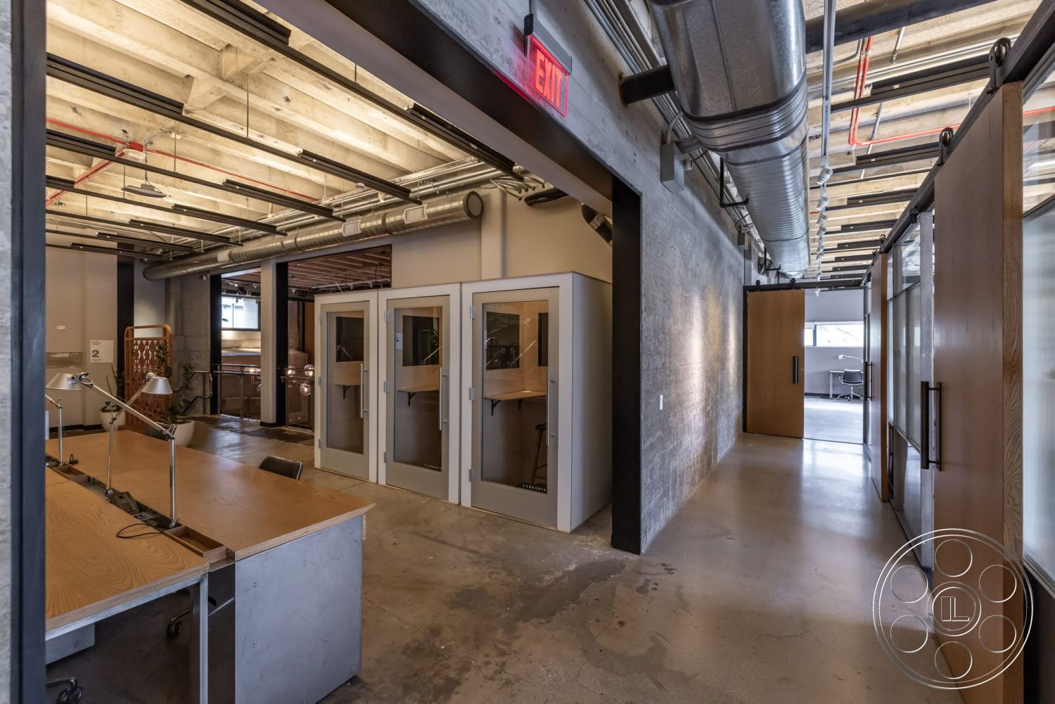 Sunset Campus - neutral color palette, polished concrete flooring, exposed concrete walls, workstation area, creative office environment, exposed ductwork ceiling, open-plan office layout, industrial style lighting, wooden table with metal legs, modern workspace interior, glass-panelled office booths, sliding wooden doors, steel-framed desk lamps