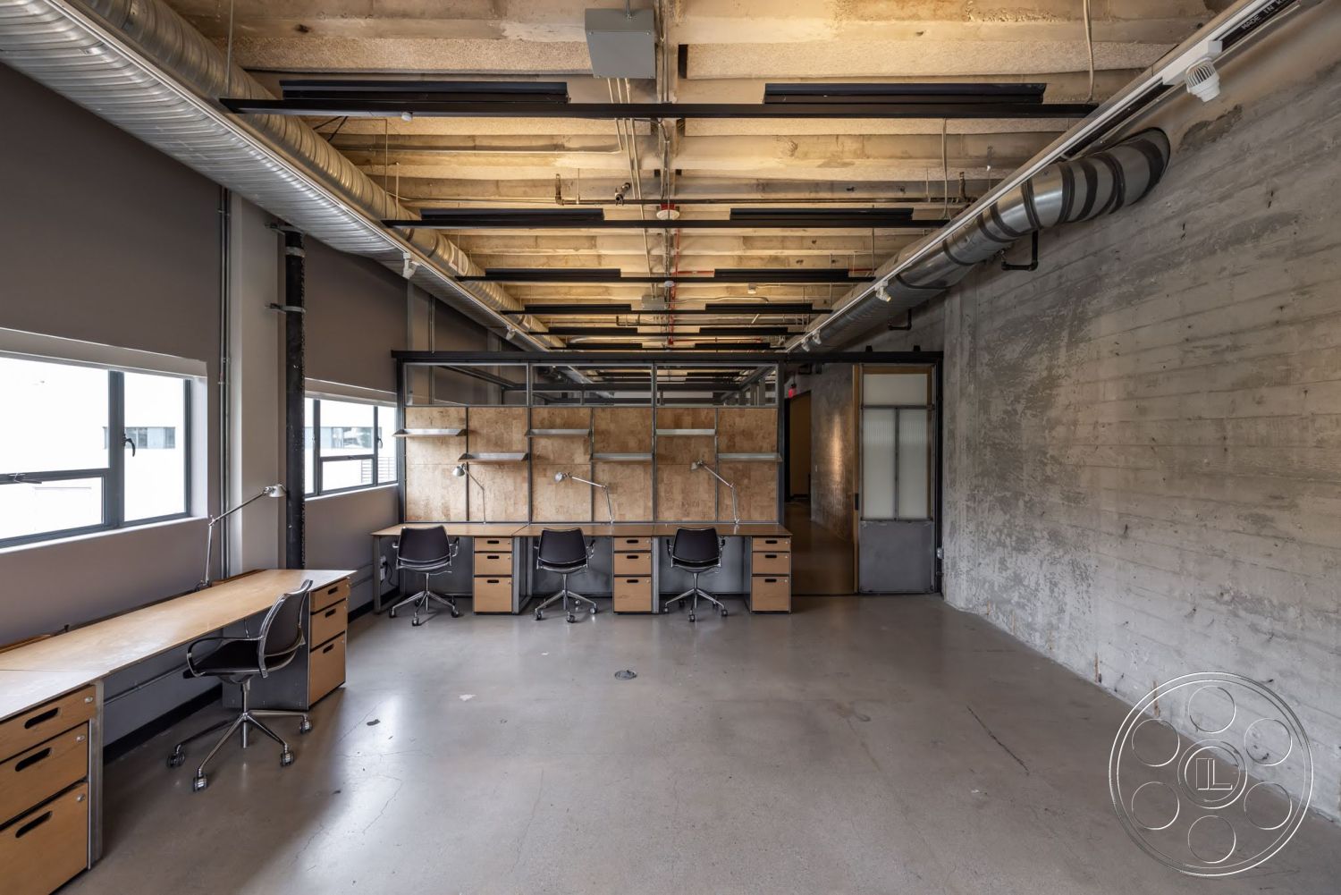 Sunset Campus - open floor plan, concrete flooring, exposed concrete walls, black office chairs, glass partition wall, large windows providing natural light, industrial-style office, metal ceiling beams, modern workspace layout, wooden desk with storage drawers, overhead ductwork