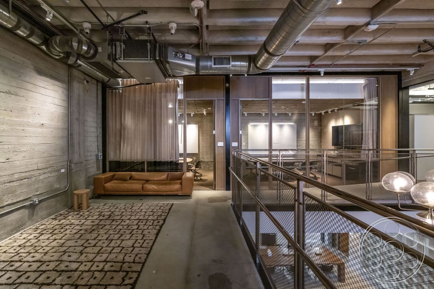 Sunset Campus - decorative area rug, concrete floor, exposed concrete walls, metal railing, track lighting, wooden accent wall, open office layout, glass partition walls, modern leather sofa, industrial ceiling ductwork