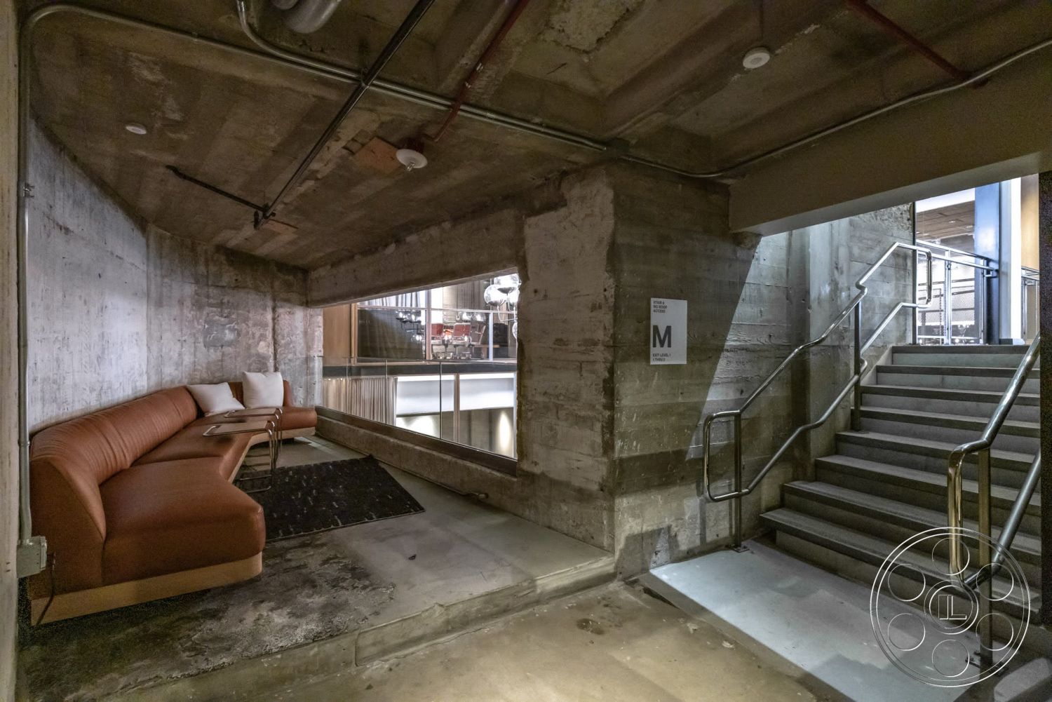 Sunset Campus - concrete flooring, open concept layout, exposed concrete walls, modern seating area, textured concrete surface, angular ceiling, metal stair railings, industrial style interior, L-shaped sectional sofa, strip lighting fixtures, staircase with straight steps, large window opening, soft furnishings with pillows