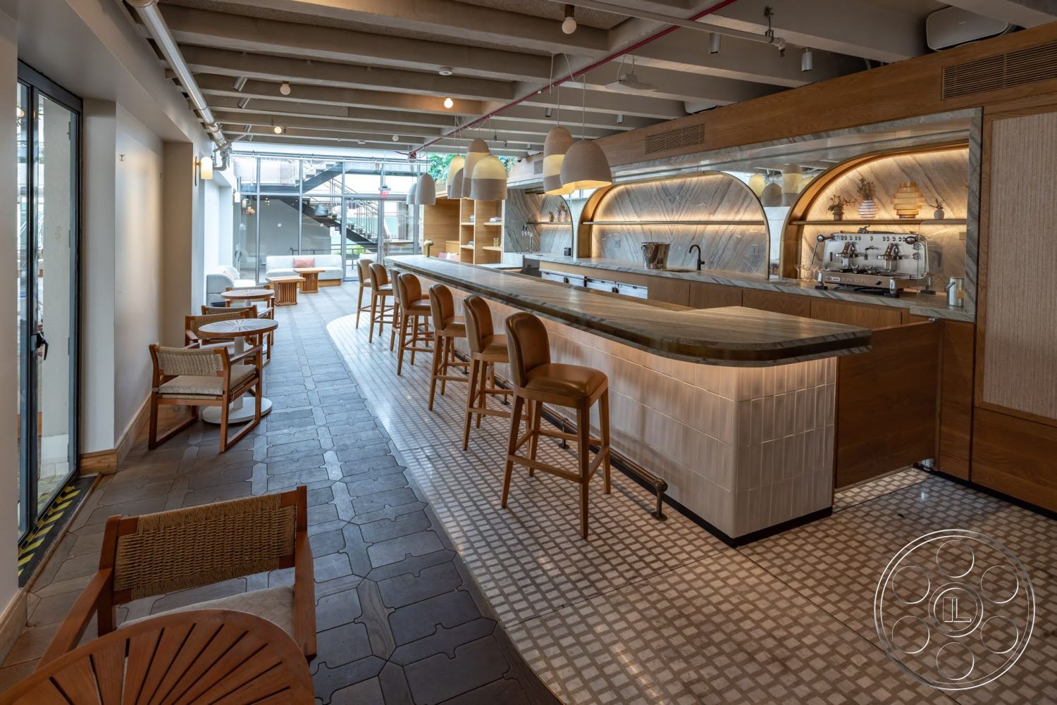 Sunset Campus - neutral color palette, natural wood cabinetry, glass sliding doors, casual dining space, soft ambient lighting, exposed concrete ceiling, wooden dining chairs, open kitchen layout, modern minimalist style, natural light emphasis, geometric patterned tile flooring, pendant light fixtures, curved bar design, wood bar countertop, white tile bar front, integrated kitchen shelving, public seating area