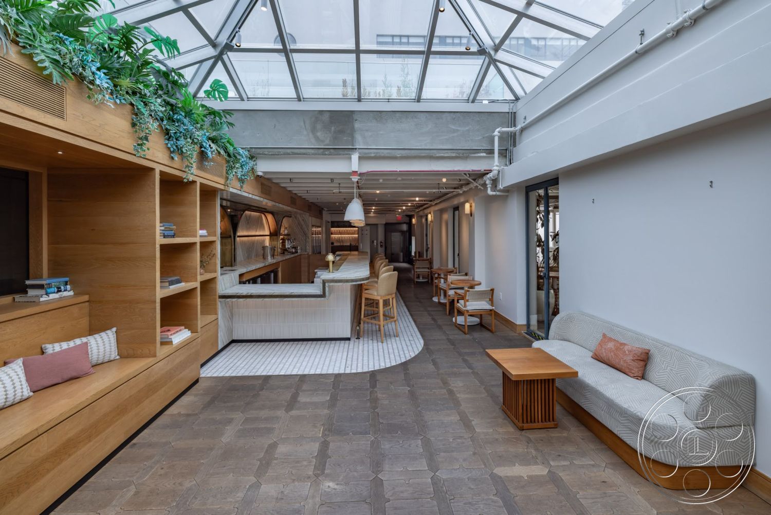 Sunset Campus - neutral color palette, indoor plant decor, open layout with natural light, concrete ceiling beams, minimalist light fixtures, hexagonal stone flooring, wooden built-in seating with bookshelves, glass roof skylights, marble kitchen bar countertop, white tile kitchen backsplash, wooden table with modern design, upholstered curved sofa with patterned fabric