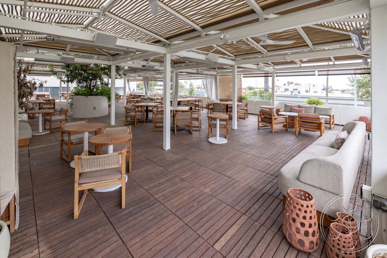 Sunset Campus - contemporary outdoor furniture, outdoor lounge area, exterior seating area, wooden deck flooring, open patio design, stainless steel light fixtures, white structural columns, woven wooden chairs, plant feature in large planter, stretched canvas awning