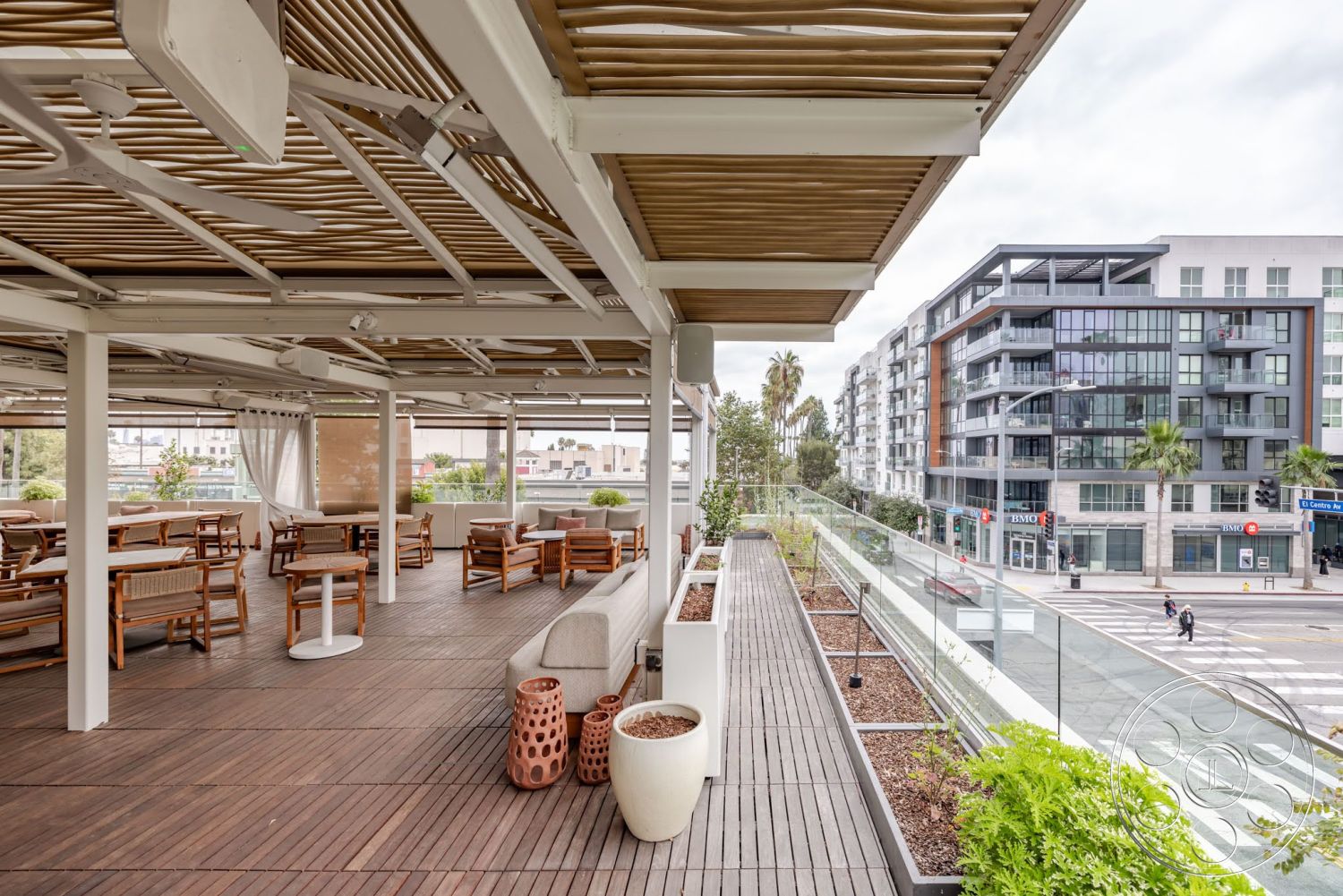 Sunset Campus - exterior, modern outdoor furniture, concrete wall, outdoor dining area, wooden deck flooring, recessed lighting fixtures, city skyline backdrop, urban setting view, planter boxes with greenery, metal railing with glass panels, slatted wood ceiling, white structural columns, fabric canopy overhead