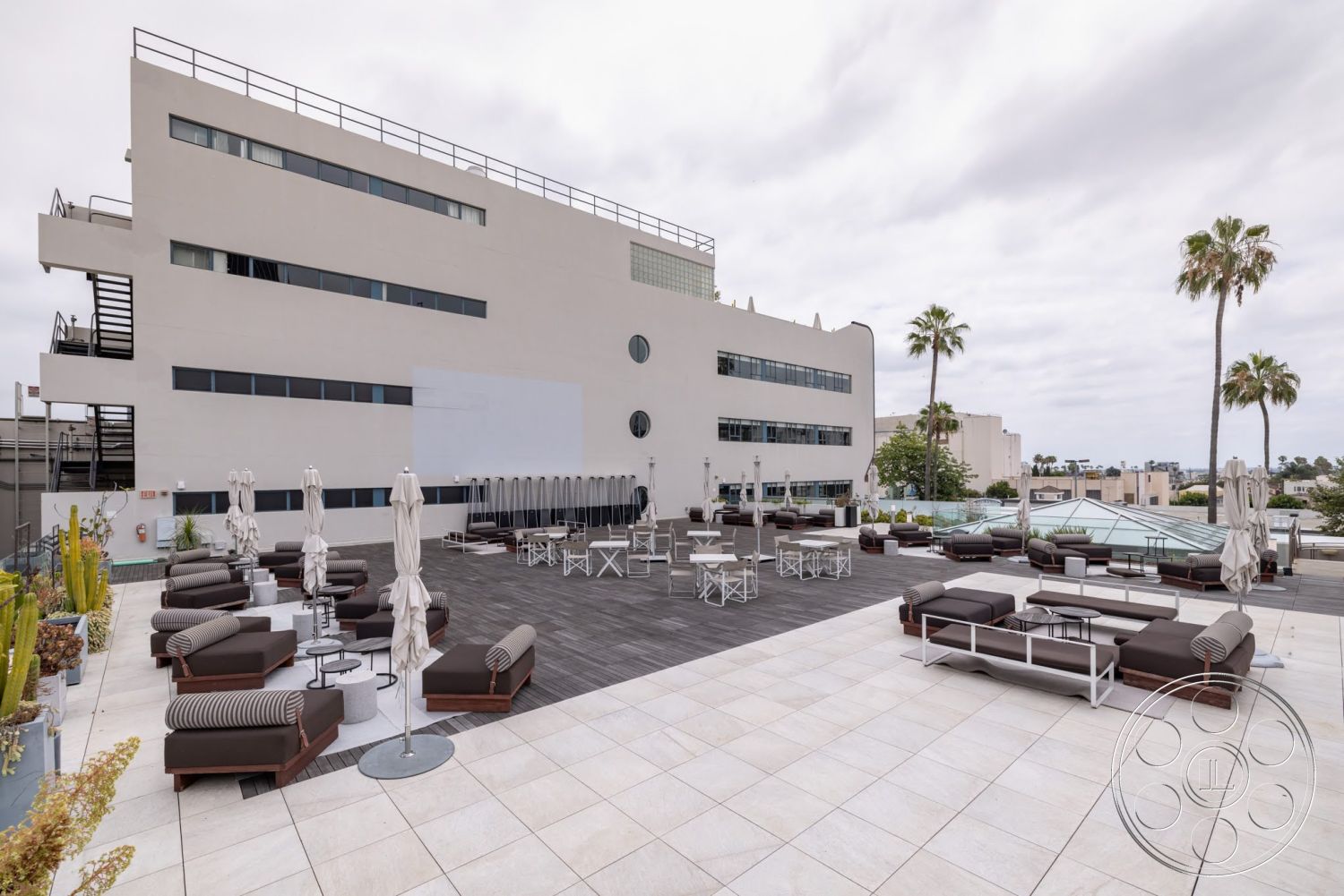Sunset Campus - exterior, floor-to-ceiling windows, gray stucco walls, wood deck flooring, planted indoor greenery, concrete terrace flooring, modern building facade, rectangular outdoor seating area, metal table and chairs, alongside palm trees, industrial staircase