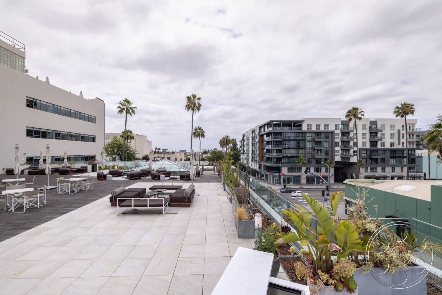Sunset Campus - exterior, glass railing, textured concrete walls, urban rooftop, city skyline view, stone patio flooring, palm trees landscaping, concrete planter boxes, wooden deck lounge chairs, rectangular outdoor lounge area, modern apartment buildings view, outdoor dining tables