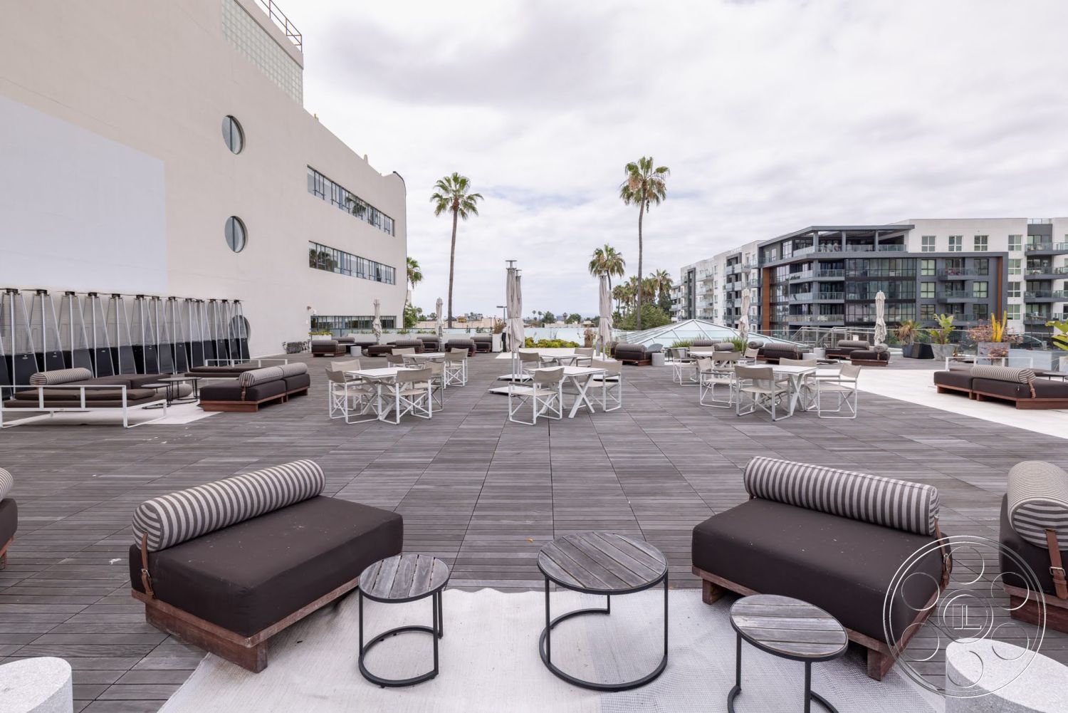 Sunset Campus - exterior, large glass windows, modern outdoor furniture, cityscape view, outdoor lounge area, sunshade umbrellas, striped outdoor cushions, concrete wall finish, minimalist lounge seating, rectangular seating arrangement, urban rooftop terrace, landscaped palm trees, composite wood decking, metal outdoor tables