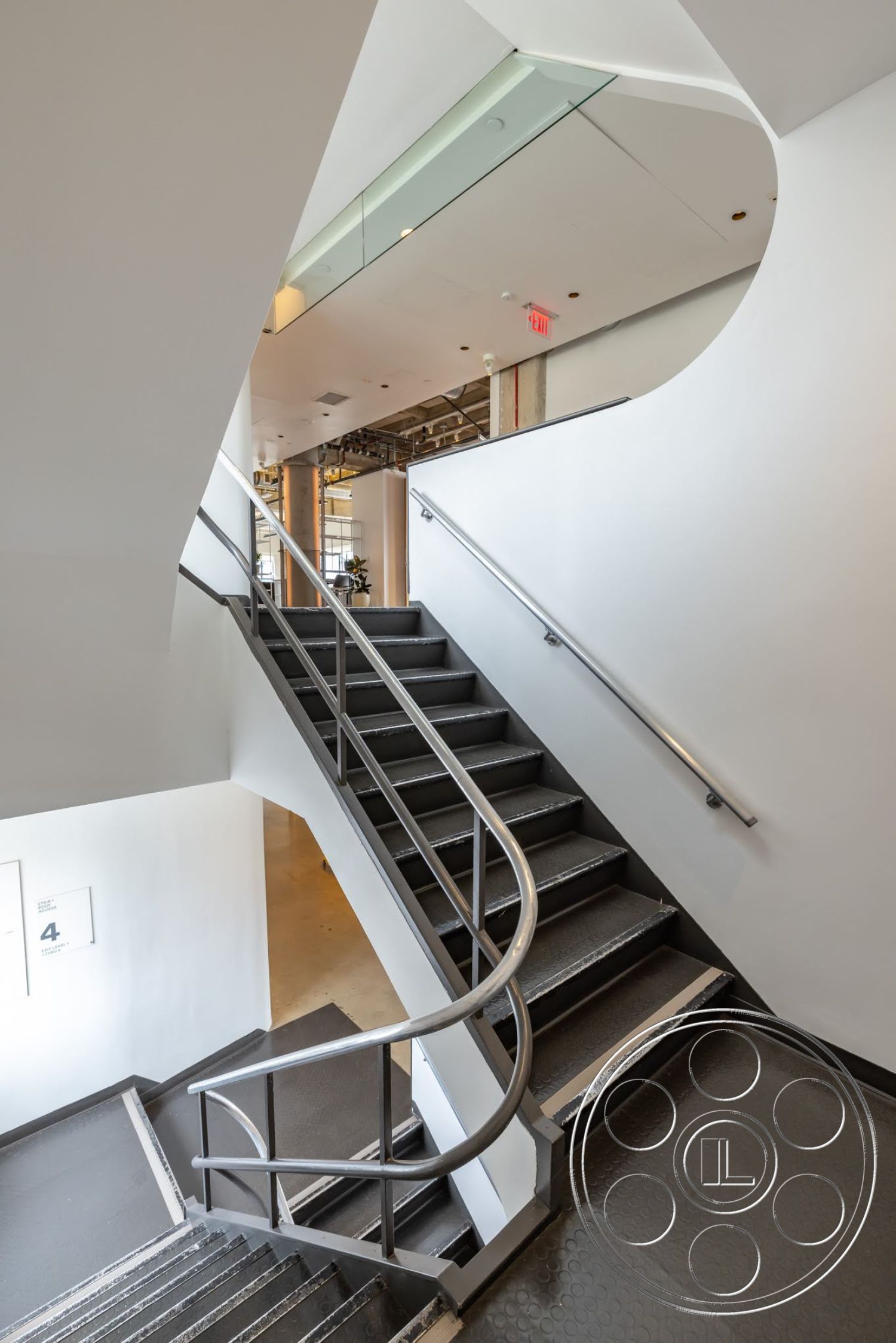 Sunset Campus - open floor plan, curved staircase, modern minimalist design, white painted walls, metal stair railings, exposed concrete structural elements, interior stairwell, textured rubber flooring, glass panel wall