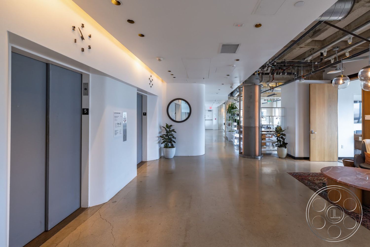 Sunset Campus - polished concrete flooring, minimalist design elements, glass partition, elevator doors, interior greenery, white painted walls, exposed ductwork ceiling, soft seating area, open layout hallway, modern round mirror, stainless steel structural columns, round decorative area rug, brushed metal light fixtures, warm wood accent details