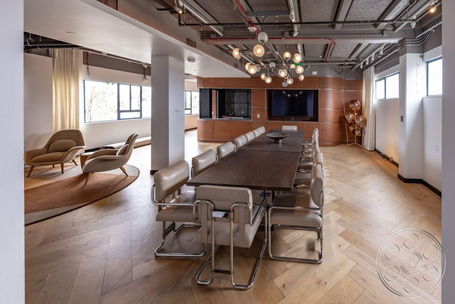 Sunset Campus - open floor plan, floor-to-ceiling windows, large flat-screen TV, circular area rug, modern minimalist design, chrome and leather dining chairs, wood panel accent wall, soft beige curtains, herringbone hardwood dining room flooring, long rectangular wooden dining table, multicolored pendant lighting fixtures, rounded lounge chairs, industrial ceiling pipes