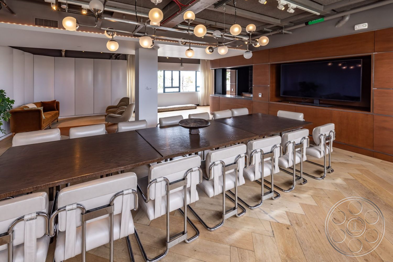 Sunset Campus - modern pendant lighting, floor-to-ceiling windows, open-concept living space, exposed concrete ceiling, hardwood herringbone dining room flooring, dark wood conference table, metal dining chairs with white upholstery, built-in wooden media console, white sheer window drapes
