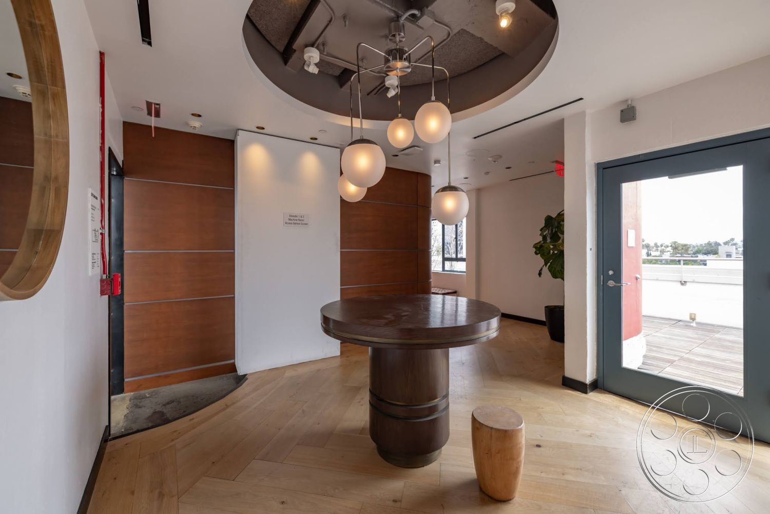 Sunset Campus - light wood flooring, white textured walls, wood panel accent wall, exposed ceiling with recessed lighting, circular wood dining table, glass patio door, pendant light installation, rounded edges in design, Indoor potted green plant