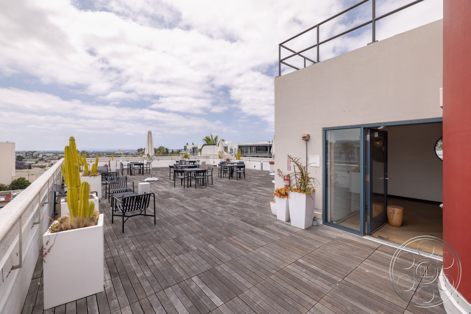 Sunset Campus - exterior, modern architecture, round table, metal railing, concrete wall, outdoor dining area, city skyline view, black metal chairs, cactus plants, white planters, umbrella shading, wooden deck flooring
