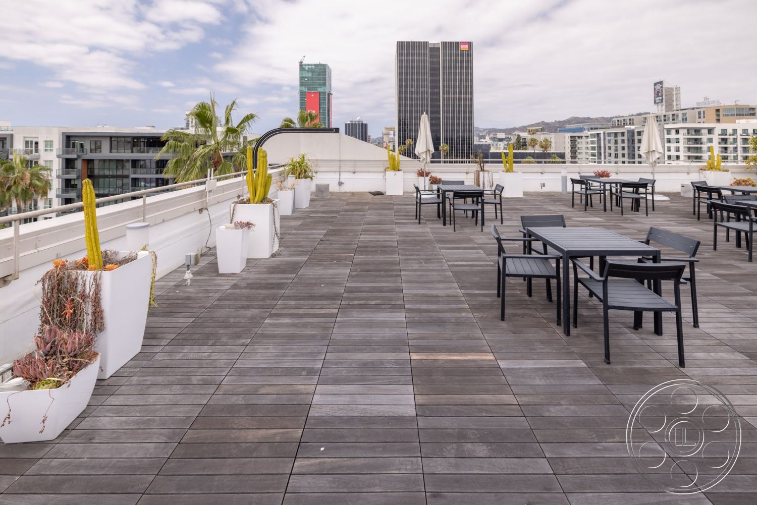 Sunset Campus - exterior, outdoor dining area, metal outdoor furniture, city skyline view, wooden deck flooring, potted cactus plants, bright sunny ambiance, contemporary rooftop terrace, planter boxes with succulents, modern building architecture