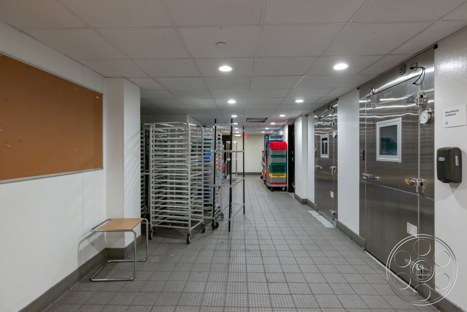 Sunset Campus - tile flooring, utility area, interior hallway, white painted walls, drop ceiling with recessed lighting, commercial kitchen environment, stainless steel freezer doors, metal storage racks, bulletin board wall feature