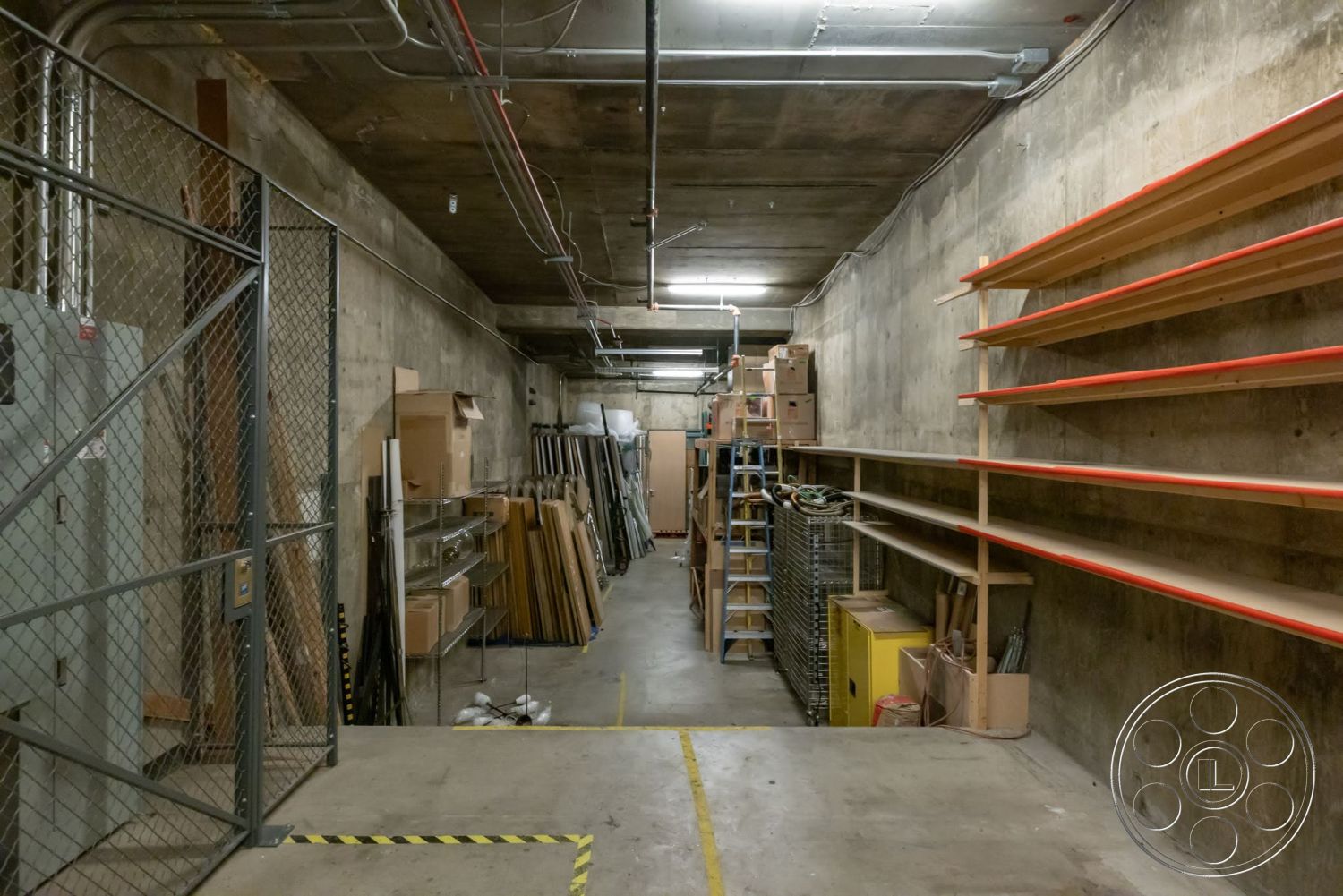 Sunset Campus - workshop area, concrete floor, exposed concrete walls, metal shelving units, storage boxes, utility room, open storage space, industrial ceiling lights, linear room layout, metal chain-link fence, wood shelving with orange trim