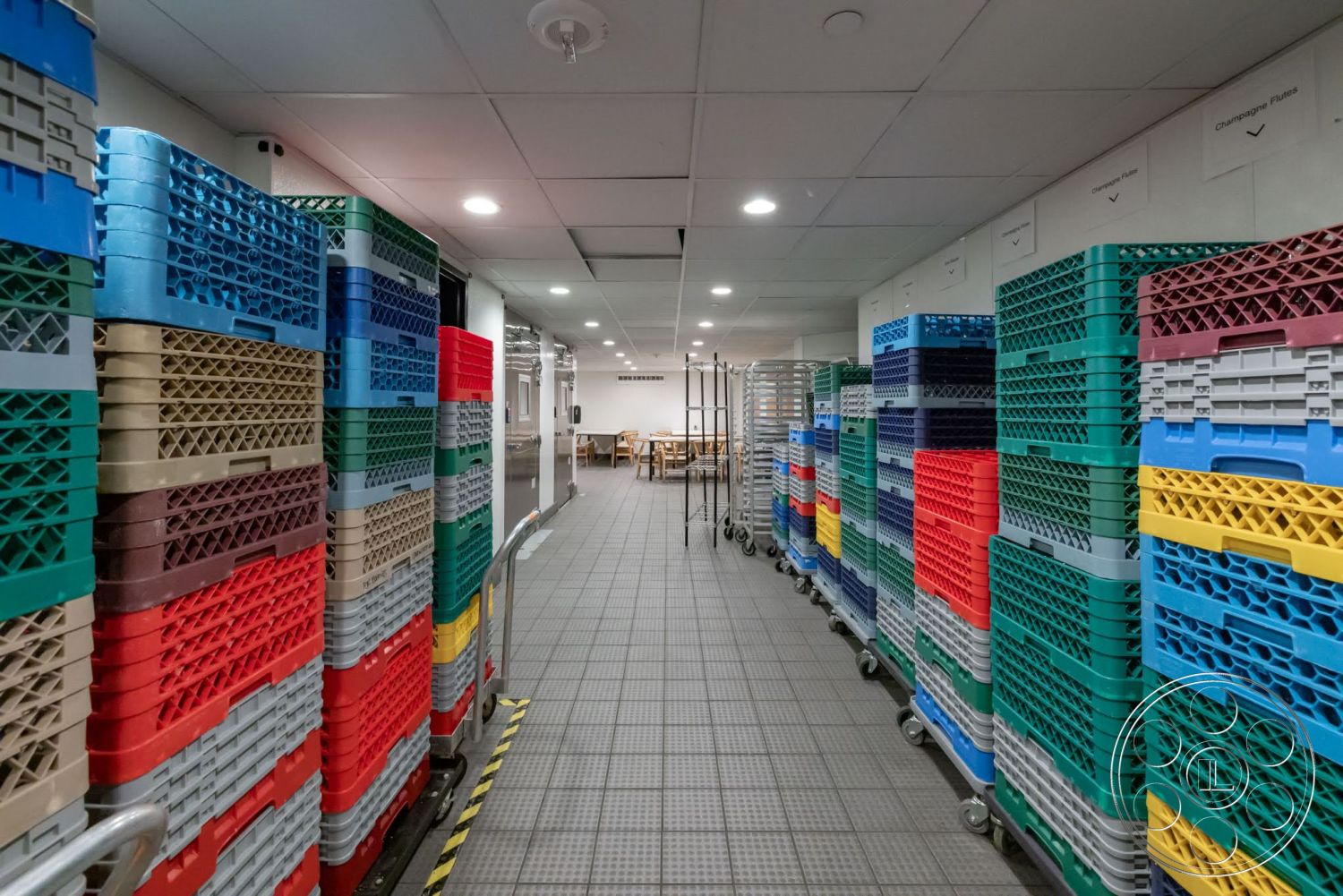 Sunset Campus - light-colored walls, low-profile ceiling, narrow hallway, bright overhead lighting, grey tiled flooring, plastic storage containers, stacked plastic crates, metal carts