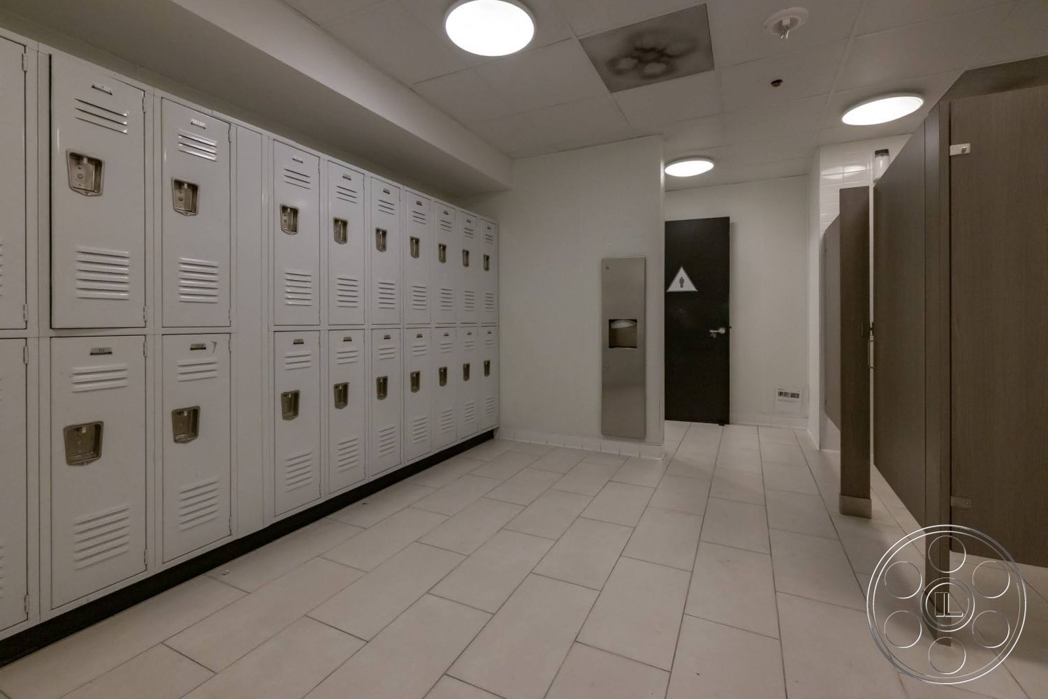 Sunset Campus - tiled flooring, modern ceiling lights, interior locker room, white metal lockers, rectangular floor tiles, gray wooden bathroom stalls, black accent baseboards, dark door with restroom signage