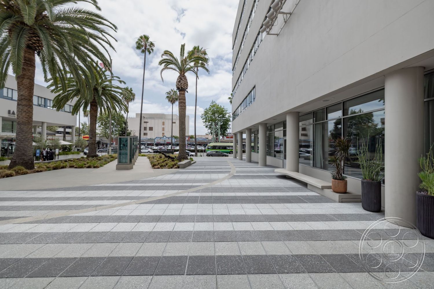 Sunset Campus - exterior, palm trees, outdoor seating area, floor-to-ceiling windows, modern architectural style, curved walkway, landscaped garden beds, commercial property setting, concrete building facade, polished concrete pavers, gray patterned paving stones, planters with decorative grasses