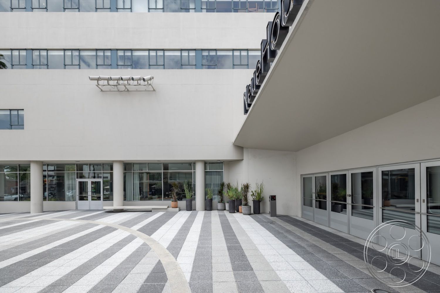 Sunset Campus - exterior, large glass windows, planters with greenery, smooth concrete walls, contemporary architectural style, metal signage, modern entryway design, curved patio layout, striped concrete patio flooring, polished stone pavers, overhanging roof, vertical natural lighting