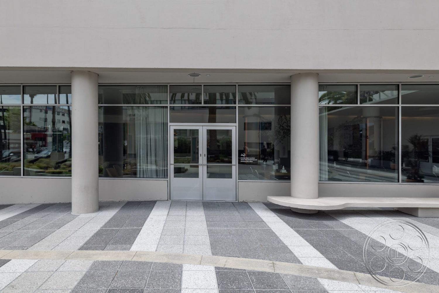 Sunset Campus - exterior, floor-to-ceiling windows, modern architectural design, glass entrance doors, concrete column supports, landscaped outdoor area, white stucco building facade, polished granite plaza flooring, dynamic geometric patterns in paving, minimalistic bench seating