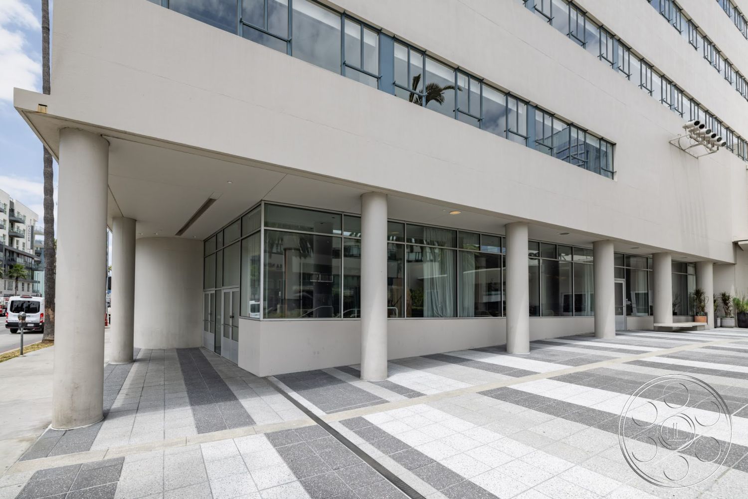 Sunset Campus - exterior, floor-to-ceiling windows, modern architectural style, textured concrete wall, rectangular patio layout, glass storefront windows, concrete column supports, palm trees in the environment, gray concrete sidewalk, checkerboard tile pavers