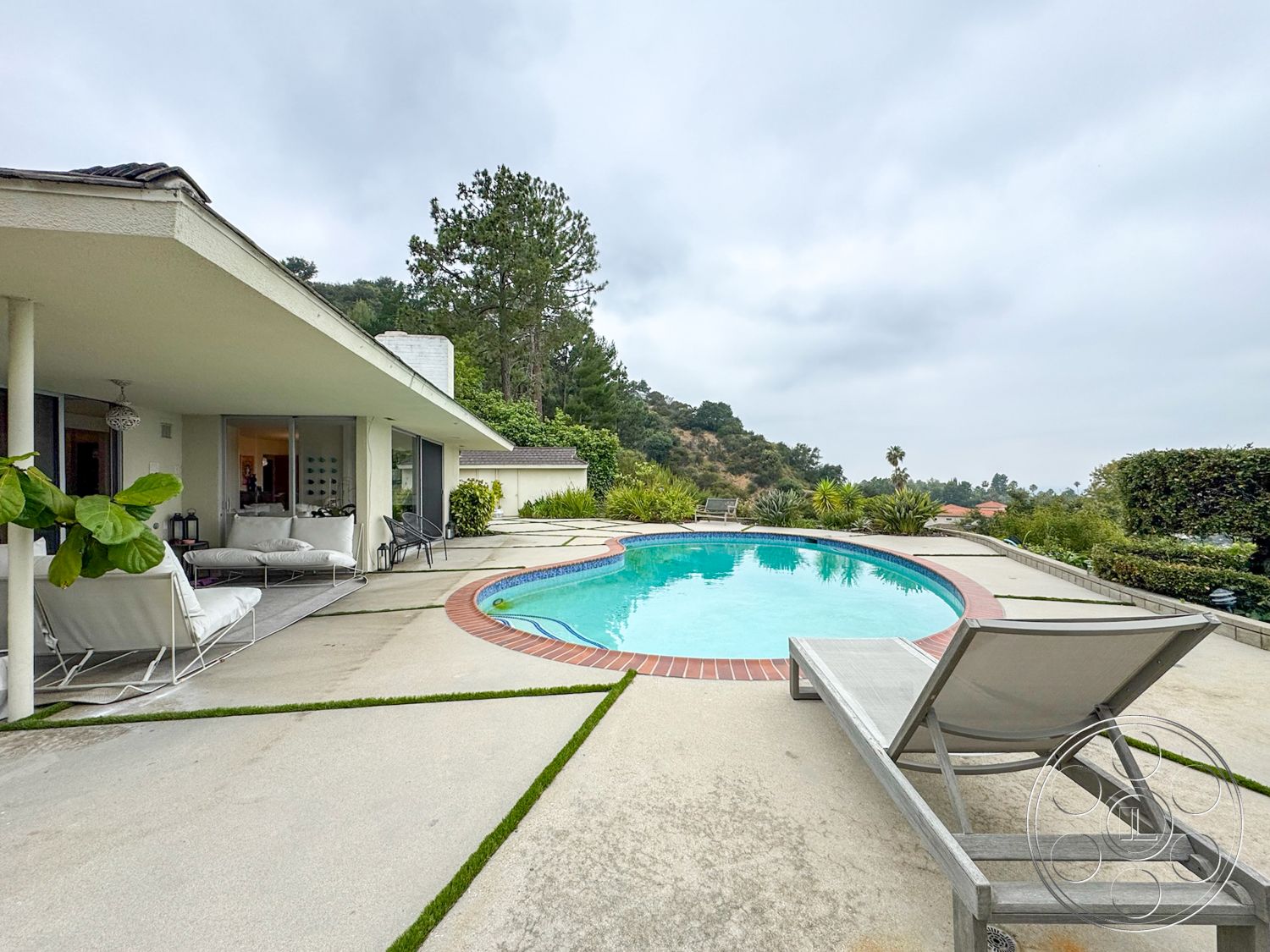 Once Upon Hollywood - exterior, glass sliding doors, outdoor seating area, contemporary architecture, planted greenery, wooden lounge chair, concrete patio flooring, rectangular pool with brick border, sloping hillside landscape, covered overhang