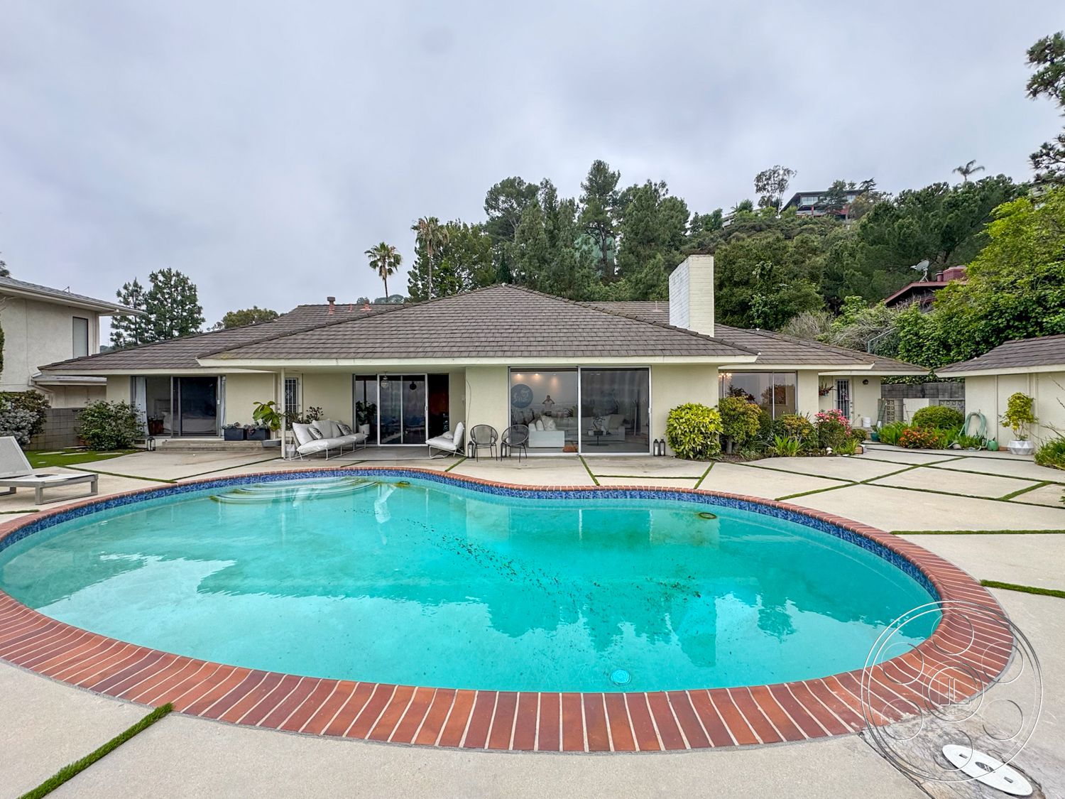 Once Upon Hollywood - exterior, lush greenery, large glass sliding doors, modern outdoor furniture, single-story home, paved walkway, rectangular pool, shingle roof, palm trees in background, landscaped garden area, concrete patio flooring, concrete pool border, brick pool coping, hilltop view setting
