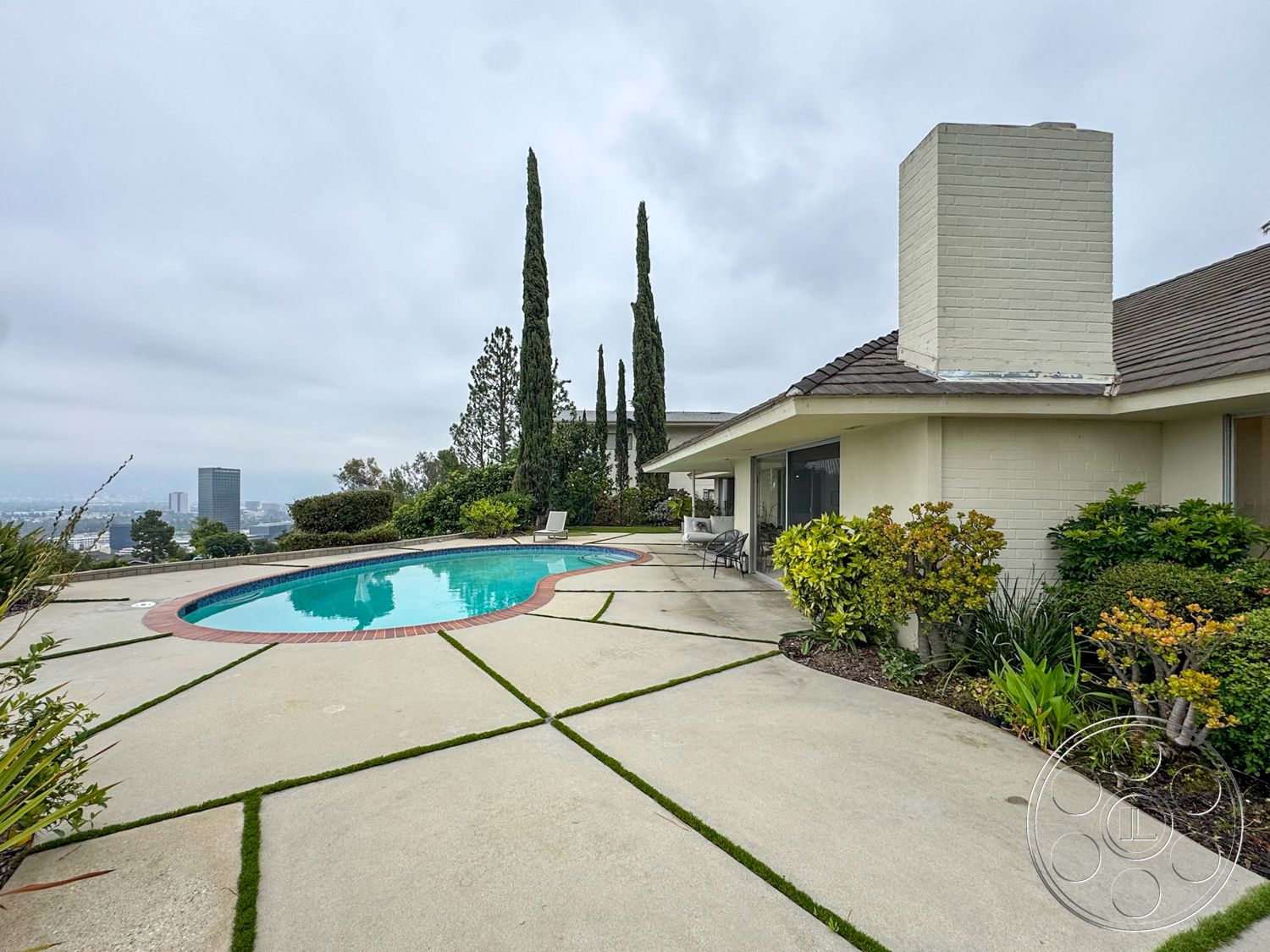 Once Upon Hollywood - exterior, large glass windows, outdoor seating area, sloped roof, single-story home, modern architectural style, city skyline view, concrete pool deck, rectangular pool, concrete patio flooring, landscaped garden beds, brick pool border, evergreen cypress trees