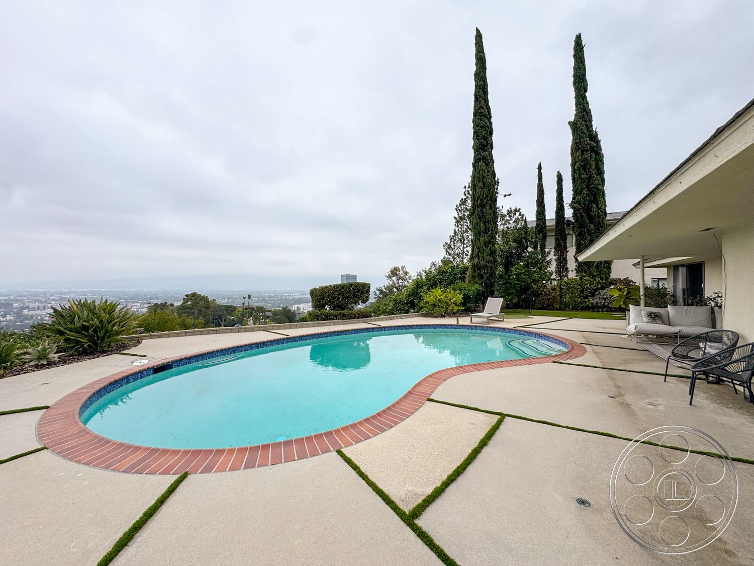 Once Upon Hollywood - exterior, tall cypress trees, concrete pool deck, landscaped garden with shrubs, brick pool border, modern house exterior, cloudy sky setting, kidney-shaped pool, concrete patio with grass strips, views of urban landscape