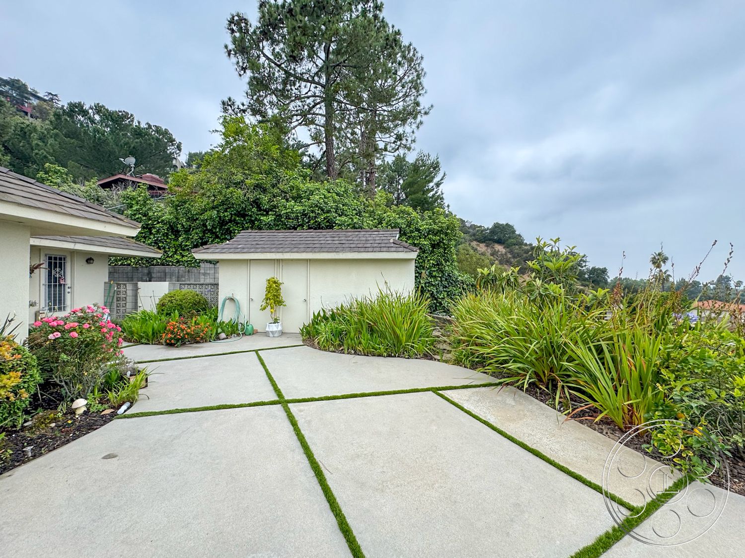 Once Upon Hollywood - single-story home, outdoor garden area, sloped hillside setting, landscaped garden with flowering plants, gabled roof structure, concrete driveway with grass insets, exterior wall treatment with stucco finish, mixed greenery including shrubs and small trees, pathway bordered by concrete and grass