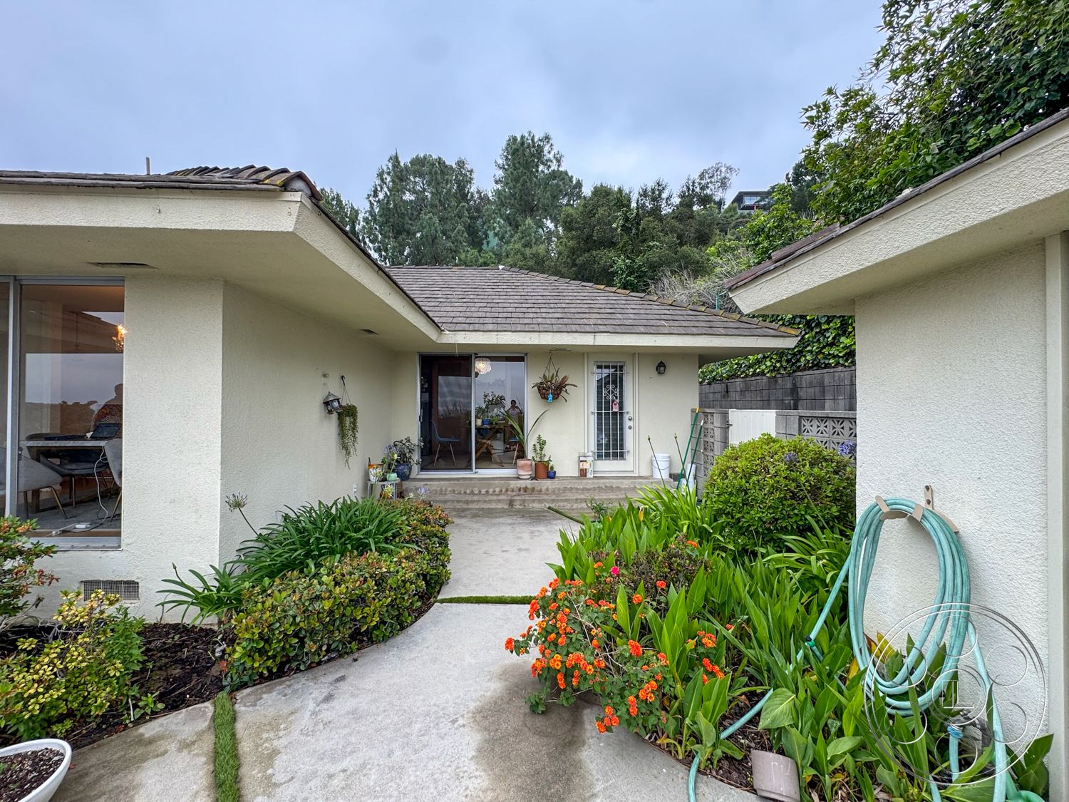 Once Upon Hollywood - exterior, green foliage, single-story home, urban garden, flowering plants, concrete walkway, decorative potted plants, wooden front door, courtyard entrance, brick retaining wall, landscaped garden beds, sloped roof with shingles, stucco house walls, light fixture at entrance, doorway with glass panel