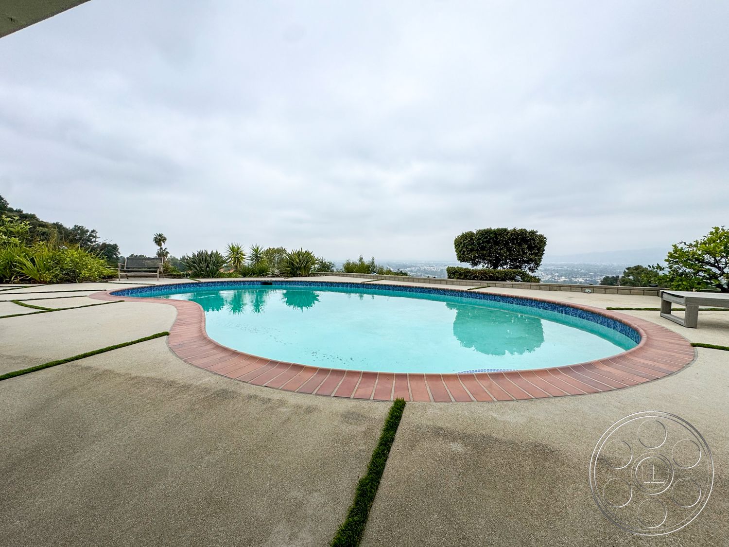 Once Upon Hollywood - exterior, smooth concrete surface, mountain view, concrete pool deck, oval pool, landscaped garden area, cloudy sky, brick pool border, grass strips between concrete, urban landscape backdrop