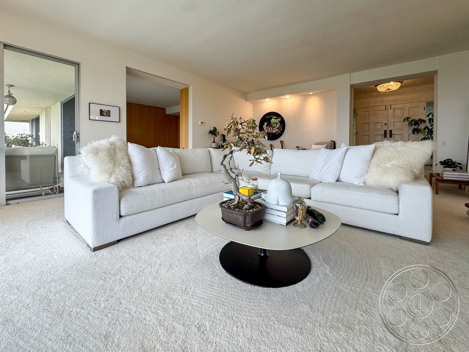Once Upon Hollywood - neutral color palette, floor-to-ceiling windows, soft lighting fixtures, decorative indoor plants, light beige carpet flooring, textured white walls, wood panel accents, bright living room, white modern sofa with throw pillows, circular coffee table with black base, access to outdoor balcony, simple artwork on wall