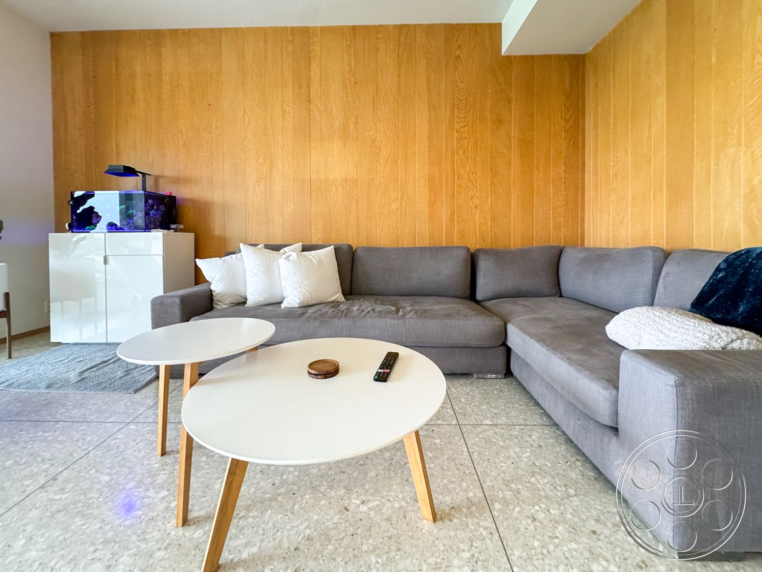 Once Upon Hollywood - minimalist decor, polished concrete flooring, bright natural lighting, grey sectional sofa, beige area rug, decorative throw pillows, wood panel accent wall, white round coffee table with wooden legs, aquarium with colorful fish