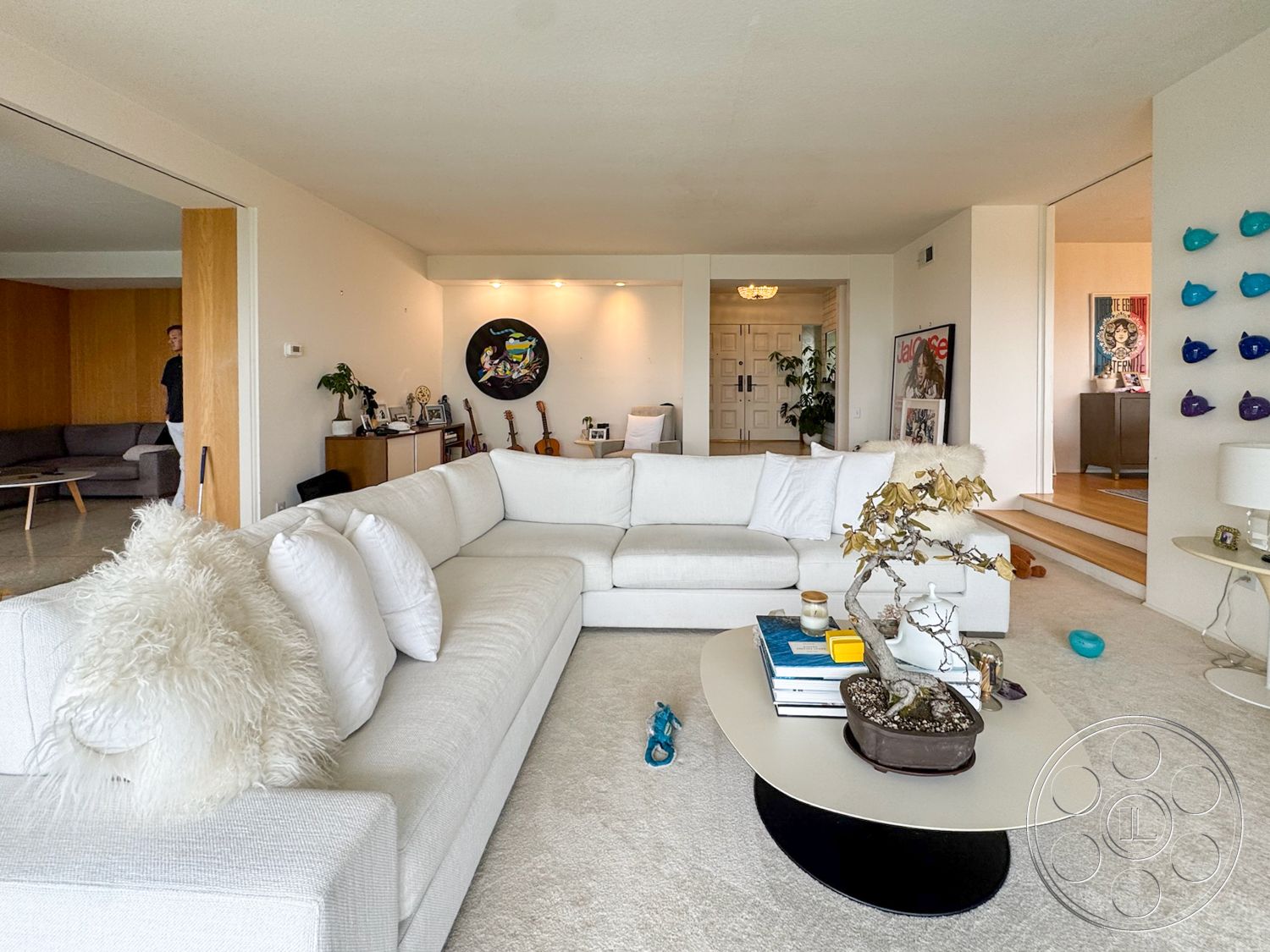 Once Upon Hollywood - round coffee table, open floor plan, artwork on walls, floor-to-ceiling windows, casual seating area, wooden side table, white sectional sofa, subtle color palette, natural light ambiance, exposed wood accent wall, modern living room, cream carpet flooring, decorative plant accent
