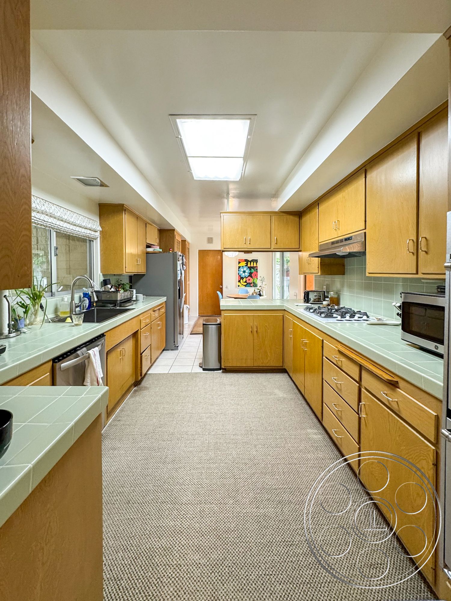 Once Upon Hollywood - stainless steel appliances, light wood cabinetry, under-cabinet lighting, open kitchen layout, interior kitchen, large window for natural light, green tile countertops, white ceiling with integrated lighting, textured beige carpet flooring, white tiled kitchen floor, fridge with side-by-side doors, gas cooktop with vent hood, kitchen sink with dual basin, colorful artwork window view, built-in cabinet storage