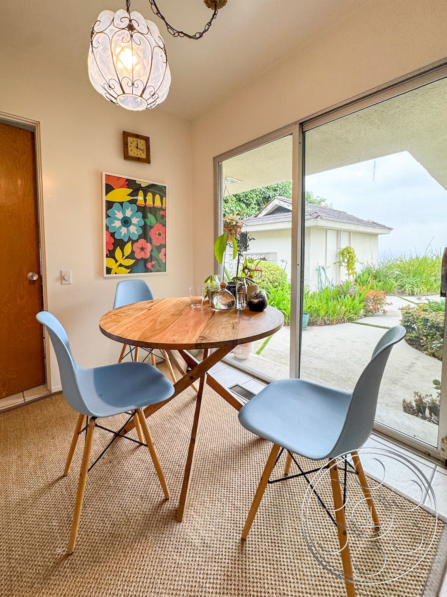 Once Upon Hollywood - indoor plants, wood dining table, glass sliding doors, painted white walls, brown wooden door, woven jute area rug, framed floral artwork, natural light dining area, blue molded plastic dining chairs, metal chandelier light fixture, siding exterior view through glass doors