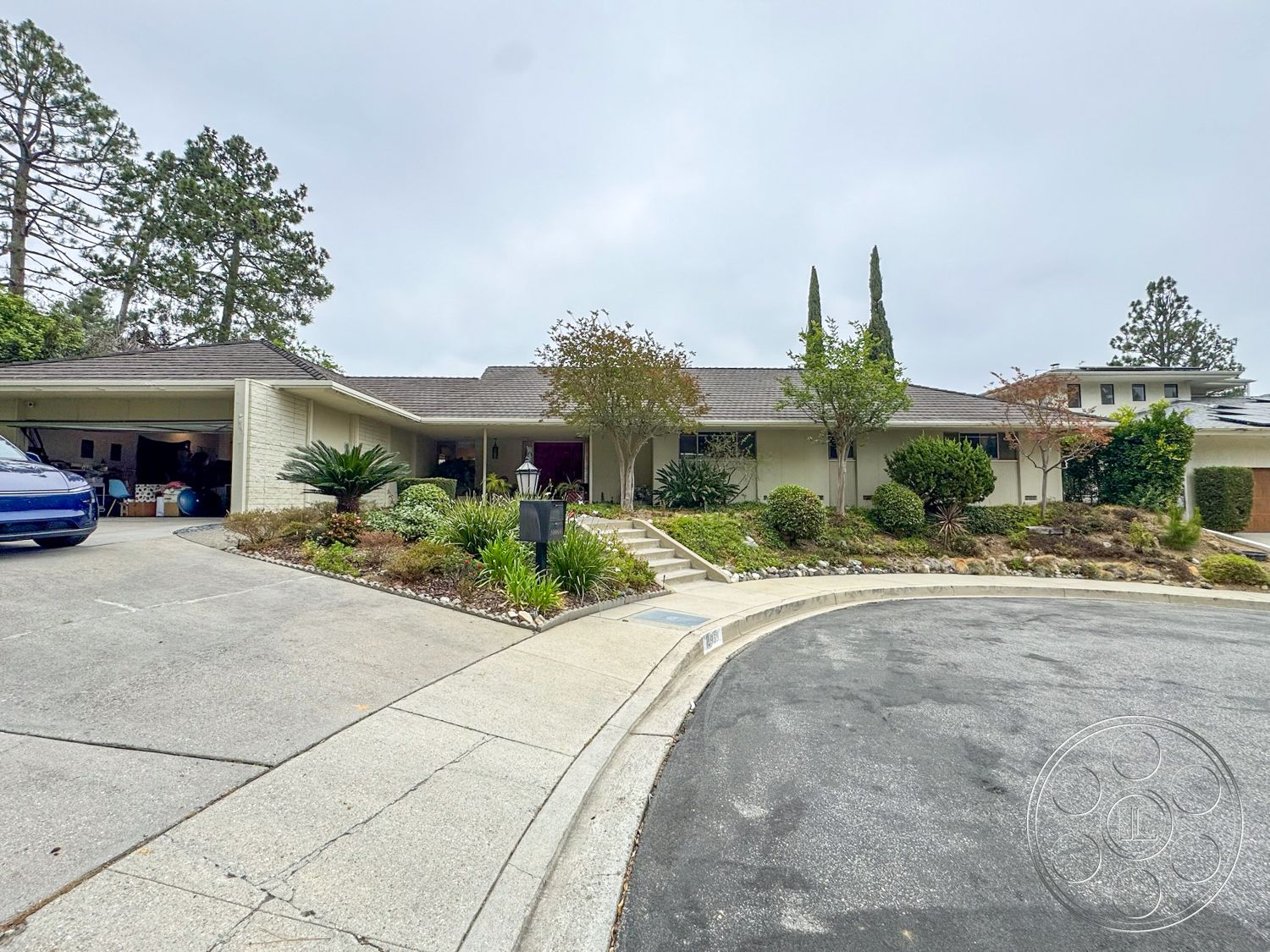 Once Upon Hollywood - exterior, asphalt shingle roof, suburban setting, decorative lanterns, planted shrubs, tall cypress trees, light-colored brick walls, single-story ranch-style home, landscaped garden beds, curved concrete driveway, rounded concrete steps, stone accent border, driveway car parking