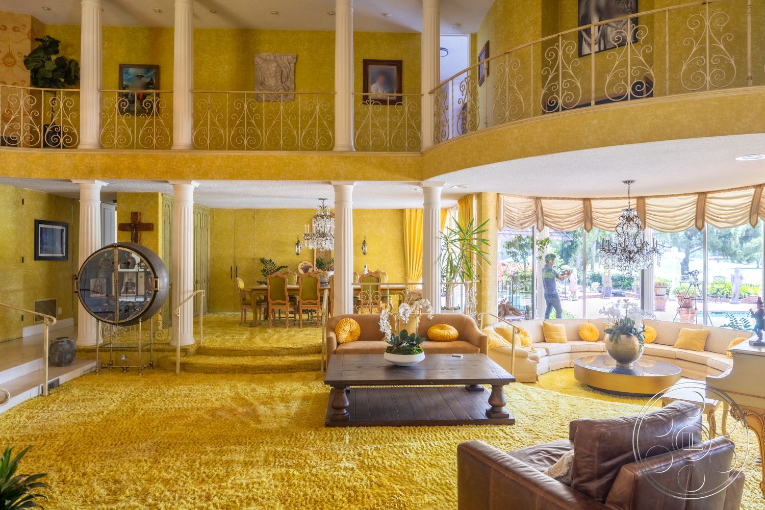 Euphoria - circular coffee table, open living space, decorative chandelier, wall-mounted artwork, exposed wood ceiling beams, modern interior design, potted indoor plants, plush carpet flooring, yellow wallpaper, sectional sofa with yellow cushions, dining area with round table and upholstered chairs, white decorative railings, large windows with drapery, round pillar supports