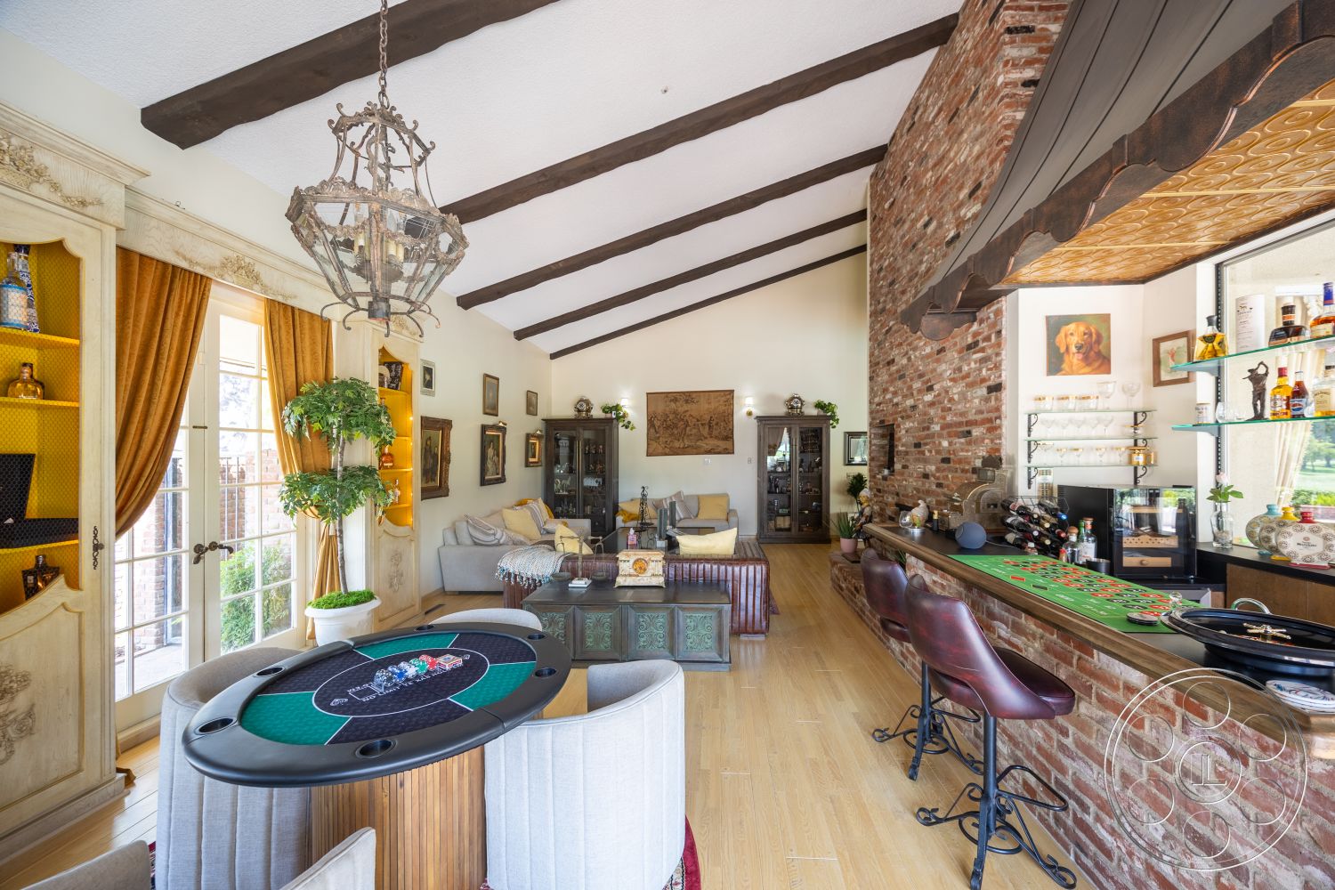Euphoria - hardwood flooring, upholstered chairs, green potted plant, exposed wood ceiling beams, vaulted living room ceiling, exposed brick accent wall, framed artwork on walls, decorative chandelier light fixture, poker table with felt top, glass bar shelving, curtains with gold accents, wood cabinet with glass doors