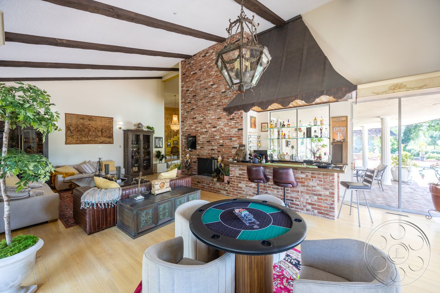 Euphoria - decorative area rug, stone fireplace, open floor plan, indoor greenery, bar stools, plush seating area, large sliding glass doors, red brick accent wall, exposed wood ceiling beams, framed artwork on wall, light hardwood living room floors, glass bar shelving, traditional lantern light fixture, game table, contemporary furniture elements