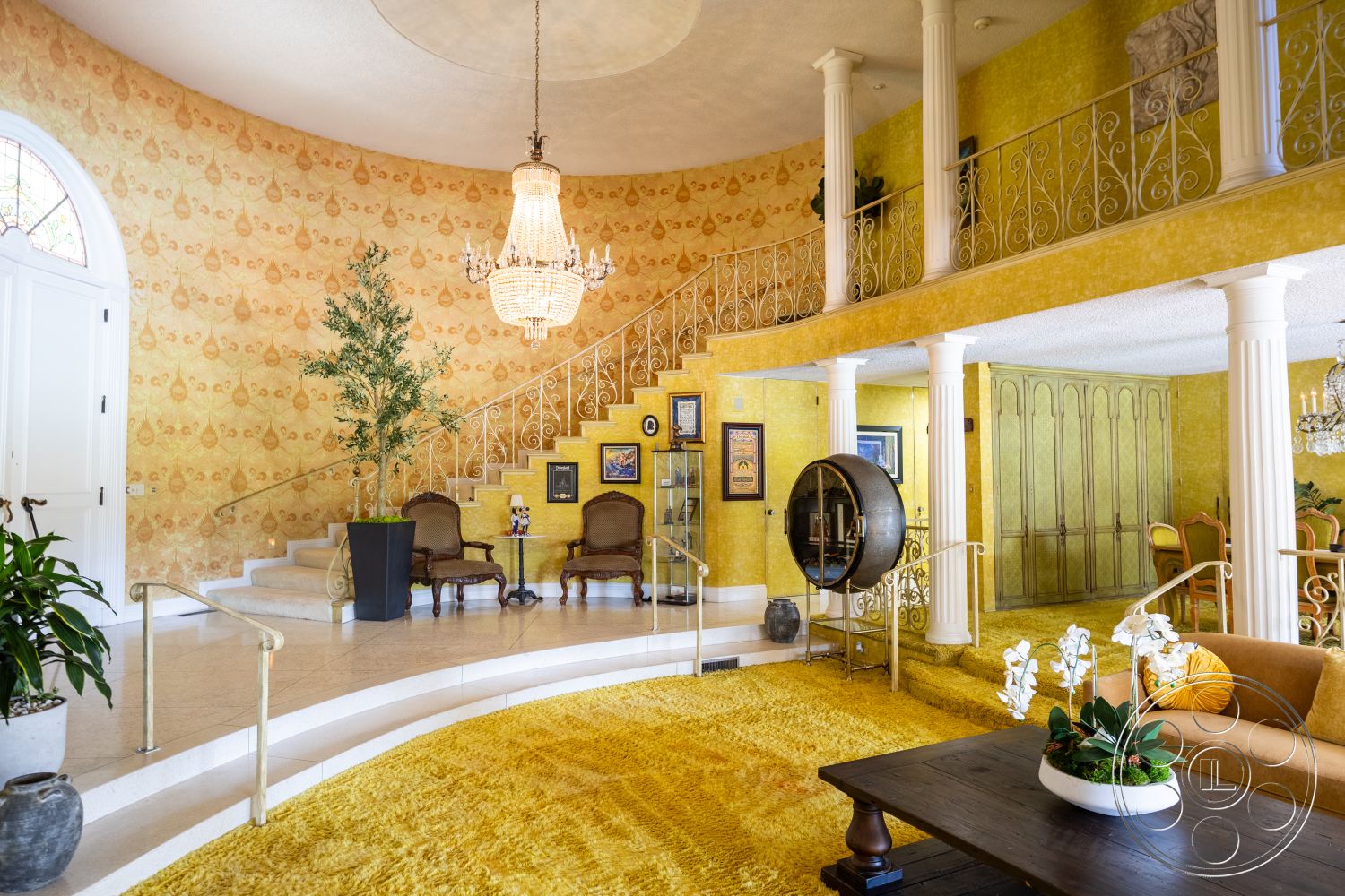 Euphoria - round coffee table, dark wood furniture, decorative wall art, exposed ceiling, arched doorways, textured plaster walls, glass display cabinet, green potted plant, curved staircase, interior foyer, white chandelier, white marble steps, yellow floral wallpaper, cream armchairs, bright yellow area rug, brass handrail