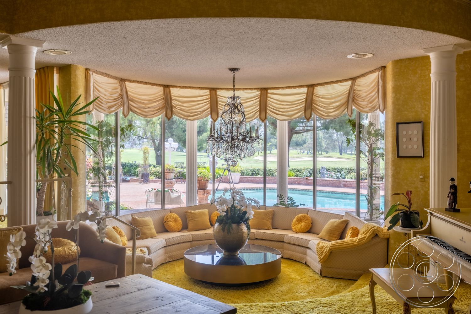 Euphoria - glass coffee table, yellow accent pillows, floor-to-ceiling windows, cozy living space, textured wall treatment, marble columns, bright yellow walls, chandelier light fixture, round sectional sofa, tall indoor plants, cream upholstery sofa, golden area rug, draped fabric window treatments, background swimming pool area, planted garden view