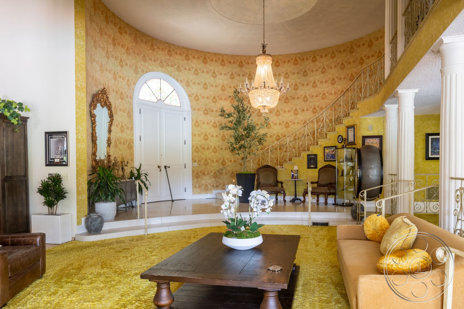 Euphoria - textured area rug, high ceiling, chandelier lighting, grand entrance, open foyer, circular staircase, exterior entrance, decorative wall mirrors, beige upholstered sofa, hardwood coffee table, classical column details, marble steps, green potted plants, yellow patterned wallpaper, white double doors, wooden accent cabinet, corner glass display case, ornate framed artwork